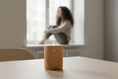 Load image into Gallery viewer, A lit wood-grain candle vessel on a table, with a softly blurred person seated near a window in the background.